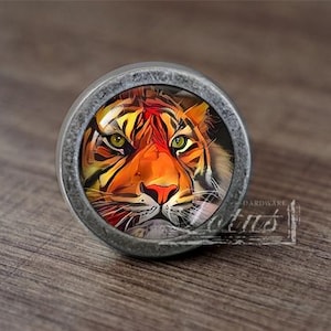 May include: A round, silver-toned cabinet knob with a tiger face design. The tiger has orange, black, and white stripes and green eyes.