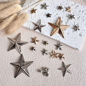 Puede incluir: Un surtido de adornos en forma de estrella en varios tamaños y acabados metálicos, incluyendo plata, oro y bronce. Las estrellas están dispersas sobre una superficie blanca texturizada, algunas sobre un cuadrado blanco.