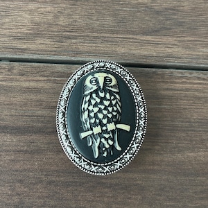 Puede incluir: Un broche de camafeo de búho blanco y negro con un marco ovalado plateado. El búho está posado en una rama y tiene un diseño detallado.