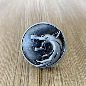 Puede incluir: Un pin de metal plateado con un diseño de cabeza de lobo. El lobo está mirando hacia la izquierda y tiene una expresión de gruñido. El pin es circular y tiene un acabado liso y pulido.