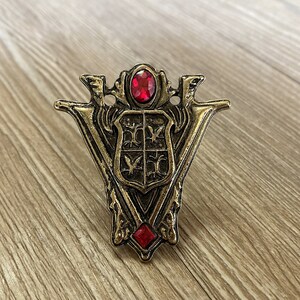 May include: A gold-colored metal pin with a red jewel in the center. The pin features a shield with a stylized design and two swords crossed behind it.