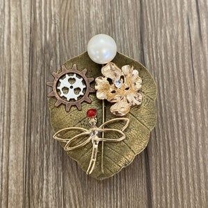 May include: A gold-toned brooch featuring a dragonfly, a flower, a gear, and a pearl. The brooch is set on a leaf-shaped base.