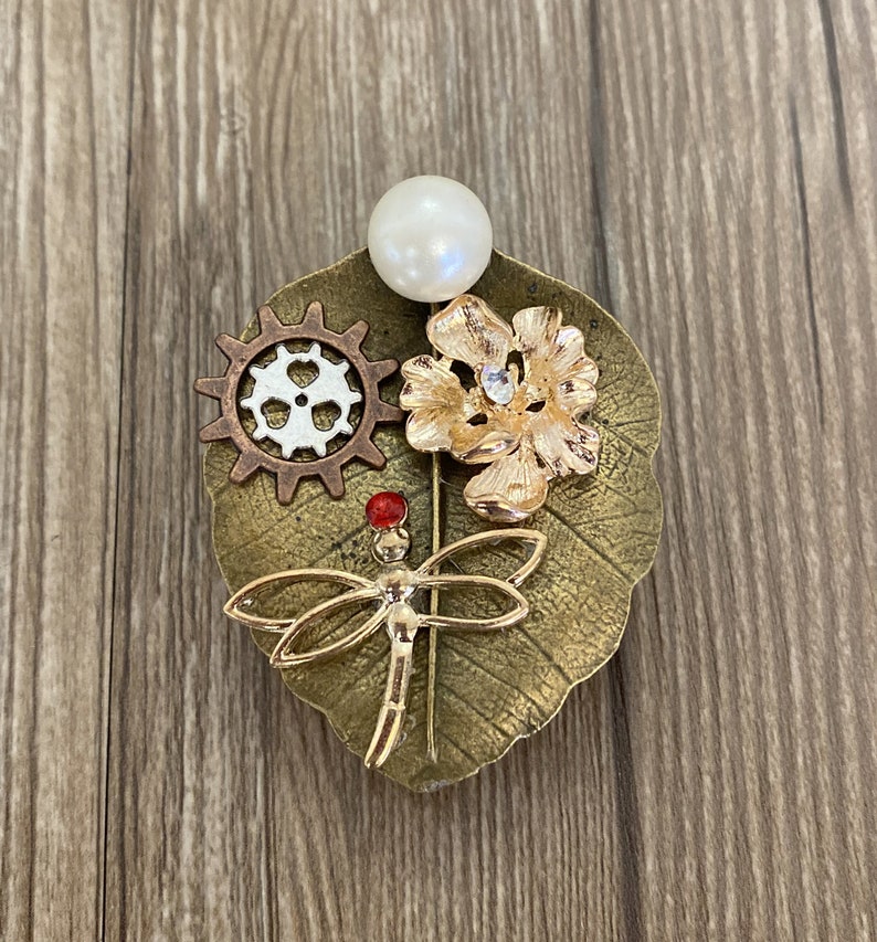 May include: A gold-toned brooch featuring a dragonfly, a flower, a gear, and a pearl. The brooch is set on a leaf-shaped base.
