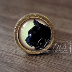 Pode incluir: Um botão de armário redondo de cor latão com uma imagem de silhueta de gato preto no centro. O gato está olhando para a esquerda e tem uma mancha branca no peito.