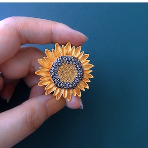 May include: A gold-toned sunflower brooch with a black rhinestone center. The sunflower has a yellow center and black petals.