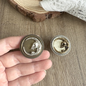 May include: Two circular metal pendants with an antique finish. Each pendant showcases a crescent moon design with a skull at its centre. The pendants have silver and bronze tones, with a beaded edge.