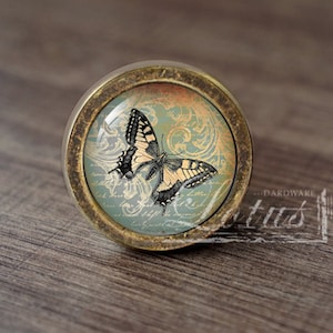 May include: A round, brass-coloured cabinet knob with a glass insert featuring a black and yellow butterfly on a blue and gold patterned background.