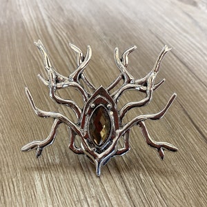 Peut inclure: Broche en argent avec un cristal brun au centre, entouré d'un motif de branches.