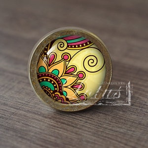 May include: A round, brass cabinet knob with a colourful, patterned glass insert. The pattern features swirls, flowers, and geometric shapes in shades of yellow, pink, green, and brown.