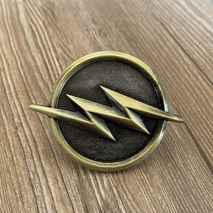 May include: A gold-coloured metal pin with a black background and a stylised lightning bolt design.