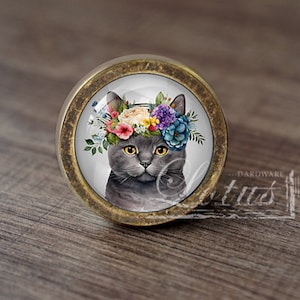 May include: A round, gold-toned cabinet knob with a glass insert featuring a grey cat wearing a colourful flower crown.