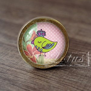 Può includere: Un pomello rotondo per armadio in ottone con un inserto in vetro che presenta un uccello verde con una corona viola. L'uccello è circondato da motivi floreali rosa e verdi.
