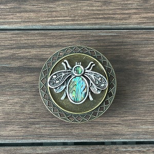 May include: A round, bronze-colored metal pin with a decorative border. The pin features a silver-colored bee with iridescent green and blue wings.