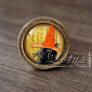 Może przedstawiać: Okrągła gałka do szafki z mosiężnym wykończeniem. Gałka przedstawia czarnego kota w pomarańczowym kapeluszu czarownicy na tle zabytkowego papieru. Dekoracyjny element okuć.