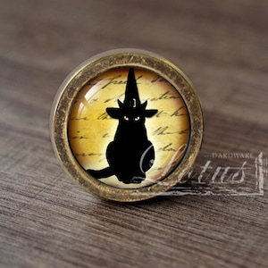 May include: A round, brass-colored cabinet knob with a glass insert featuring a black silhouette of a cat wearing a witch's hat on a yellowed, vintage-looking background with handwritten text.