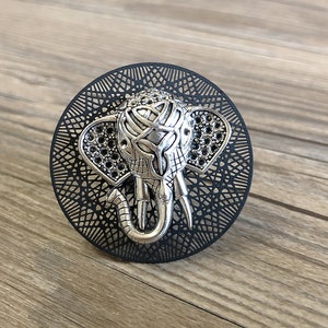 May include: A silver elephant head charm with intricate details, set against a black circular background with a geometric pattern.