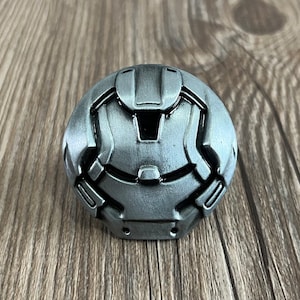 May include: A silver metal pin with a detailed design of a robot head. The robot head has a black visor and a textured surface.