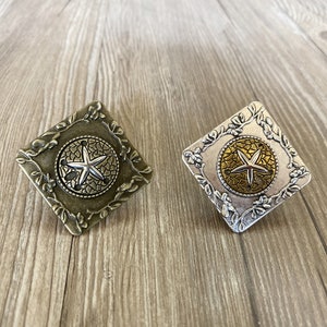 Puede incluir: Dos piezas decorativas de metal de forma cuadrada con un diseño floral y una estrella de mar dentro de un círculo en el centro. Una pieza es dorada y la otra es plateada.