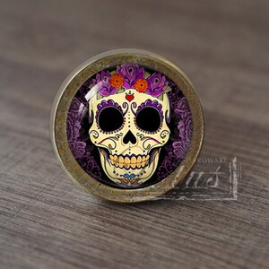 Puede incluir: Un pomo de armario redondo de color latón con un inserto de vidrio que presenta un diseño de calavera de azúcar colorida del Día de Muertos con flores moradas y acentos naranjas.