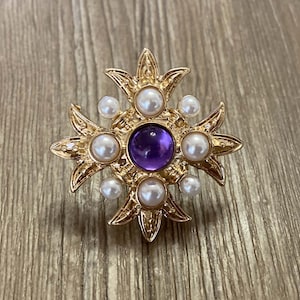 Könnte beinhalten: Eine goldfarbene Brosche mit einem violetten Edelstein in der Mitte, umgeben von weißen Perlen. Die Brosche hat die Form eines Kreuzes mit filigranen Details.