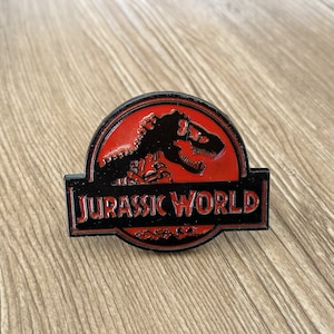 Peut inclure: Épinglette en émail noir et rouge avec le logo de Jurassic World et un squelette de dinosaure à l'intérieur du logo.