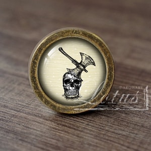 Puede incluir: Un pomo de armario redondo de color latón con un inserto de vidrio que presenta una ilustración en blanco y negro de una calavera con un hacha atravesándola.