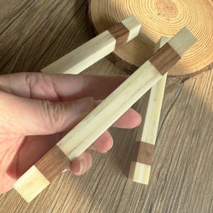 May include: Hand holding three wooden dowels. Each dowel is rectangular with a light wood body and a dark wood accent band. The dowels are on a wooden surface.