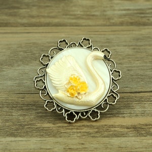 Könnte beinhalten: Eine weiße Schwanenbrosche mit gelben Blumen, eingefasst in einen silberfarbenen filigranen Rahmen.