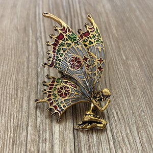 Könnte beinhalten: Eine goldfarbene Brosche mit einer Fee mit Flügeln aus Buntglas. Die Flügel sind mit roten, grünen und weißen geometrischen Mustern verziert.