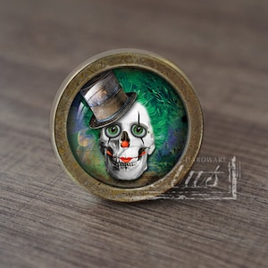 Pode incluir: Um botão de armário redondo de cor latão com uma caveira verde e branca que usa um chapéu de coco. A caveira tem um nariz vermelho e olhos verdes.