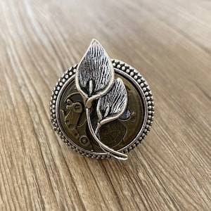 May include: A silver ring with a detailed design of two stylized leaves and a brass clockwork mechanism.