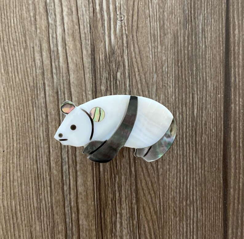 Puede incluir: Un broche de panda blanco y negro hecho de n&aacute;car. El panda tiene un ojo negro y una raya negra en la espalda.