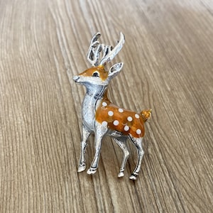 Può includere: Un broche a forma di cervo in smalto argentato e arancione con pois bianchi. Il cervo ha un collo lungo e una coda lunga. Il broche è un'ottima aggiunta a qualsiasi outfit.