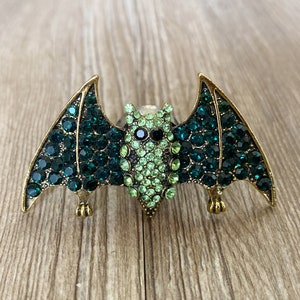 Könnte beinhalten: Eine goldfarbene Fledermaus-Brosche mit grünen und hellgrünen Strasssteinen. Die Fledermaus hat einen detaillierten Körper und Flügel.