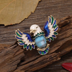 May include: A gold-toned eagle brooch with blue, green, and white enamel wings and a blue and white swirl gemstone in the center.