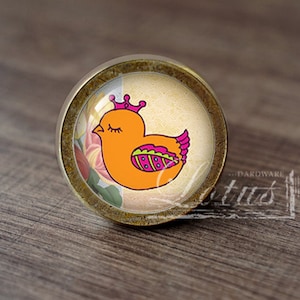 Può includere: Un pomello per armadio rotondo, color oro, con un inserto di vetro che presenta un'illustrazione di un uccello giallo con una corona rosa. L'uccello ha gli occhi chiusi ed è circondato da motivi floreali rosa e verdi.