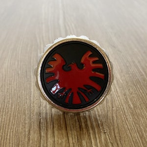 Könnte beinhalten: Eine silberfarbene Anstecknadel mit einem schwarzen und roten Adler-Design. Der Adler ist stilisiert und hat einen roten Körper und schwarze Flügel.