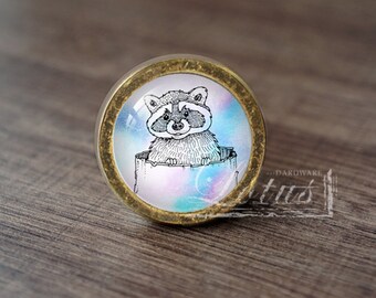 May include: A round, gold-colored cabinet knob with a glass insert featuring a black and white illustration of a raccoon peeking out of a tree trunk. The background of the glass insert is a watercolor-like design with shades of blue, pink, and purple.
