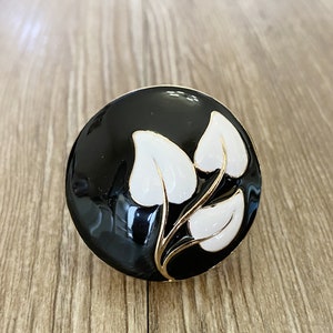 May include: A black round button with a gold trim and white floral design. The button features three stylised leaves with a gold stem.
