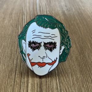 May include: A green and white enamel pin featuring a cartoon depiction of the Joker from the Batman franchise. The Joker has a white face with a red smile and green hair. His eyes are black with purple eyelids.