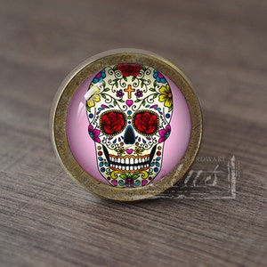 Puede incluir: Un pomo de armario redondo de color dorado antiguo con un fondo rosa y un diseño de calavera de azúcar colorido. La calavera tiene una cruz en la frente y flores alrededor de los ojos.