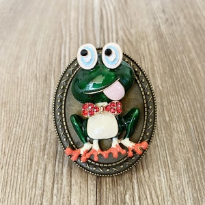 May include: A green frog with a red bow tie and white eyes is depicted on a round, bronze-coloured brooch. The frog is wearing a red bow tie with white accents and has a pink tongue sticking out.