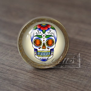 Könnte beinhalten: Ein runder, antiker goldfarbener Schrankknopf mit einem Glaseinsatz, der ein farbenfrohes Zucker Schädel-Design zeigt. Der Schädel ist weiß mit blauen, gelben, grünen und roten Details.