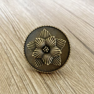 Könnte beinhalten: Ein runder, bronzefarbener Schrankknopf mit einem erhabenen, detaillierten Blumendesign.