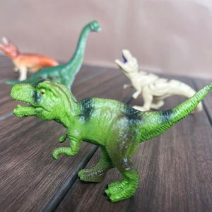 May include: A selection of four toy dinosaurs in different colours. The image shows a green Tyrannosaurus Rex with black markings. Other dinosaurs include a green Brachiosaurus, a tan dinosaur, and an orange dinosaur.