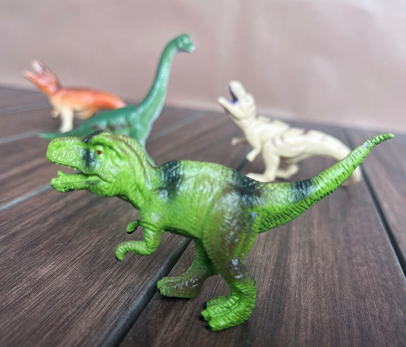May include: A selection of four toy dinosaurs in different colours. The image shows a green Tyrannosaurus Rex with black markings. Other dinosaurs include a green Brachiosaurus, a tan dinosaur, and an orange dinosaur.