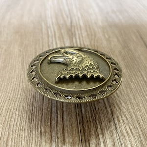 May include: A bronze-coloured eagle head ornament with a decorative border. The eagle is facing left and has a detailed design.