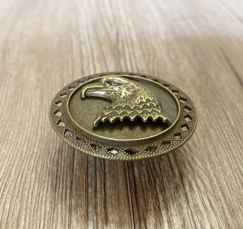 May include: A bronze-coloured eagle head ornament with a decorative border. The eagle is facing left and has a detailed design.