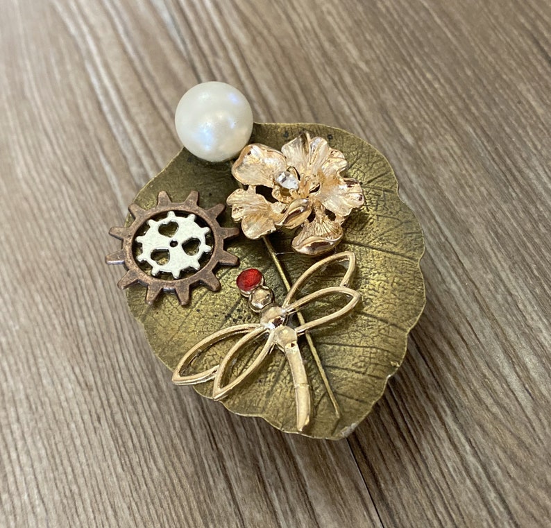 May include: A gold-toned brooch featuring a dragonfly, a flower, a gear, and a pearl. The brooch is set on a leaf-shaped base.