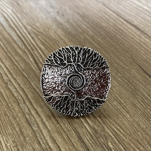Könnte beinhalten: Ein silberfarbener, runder Ring mit einem Lebensbaum-Design. Der Ring zeigt einen detaillierten Baum mit Ästen und Wurzeln sowie eine Spirale in der Mitte. Der Ring ist aus Metall und hat eine strukturierte Oberfläche.